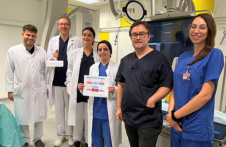 Team der Kardiologie der UMM in einem Herzkatheterlabor. Eine Mitarbeiterin hält eine Übersicht zur Studie, ein Kollege ein Modell mit der Aufschrift „Frauenherzen“.